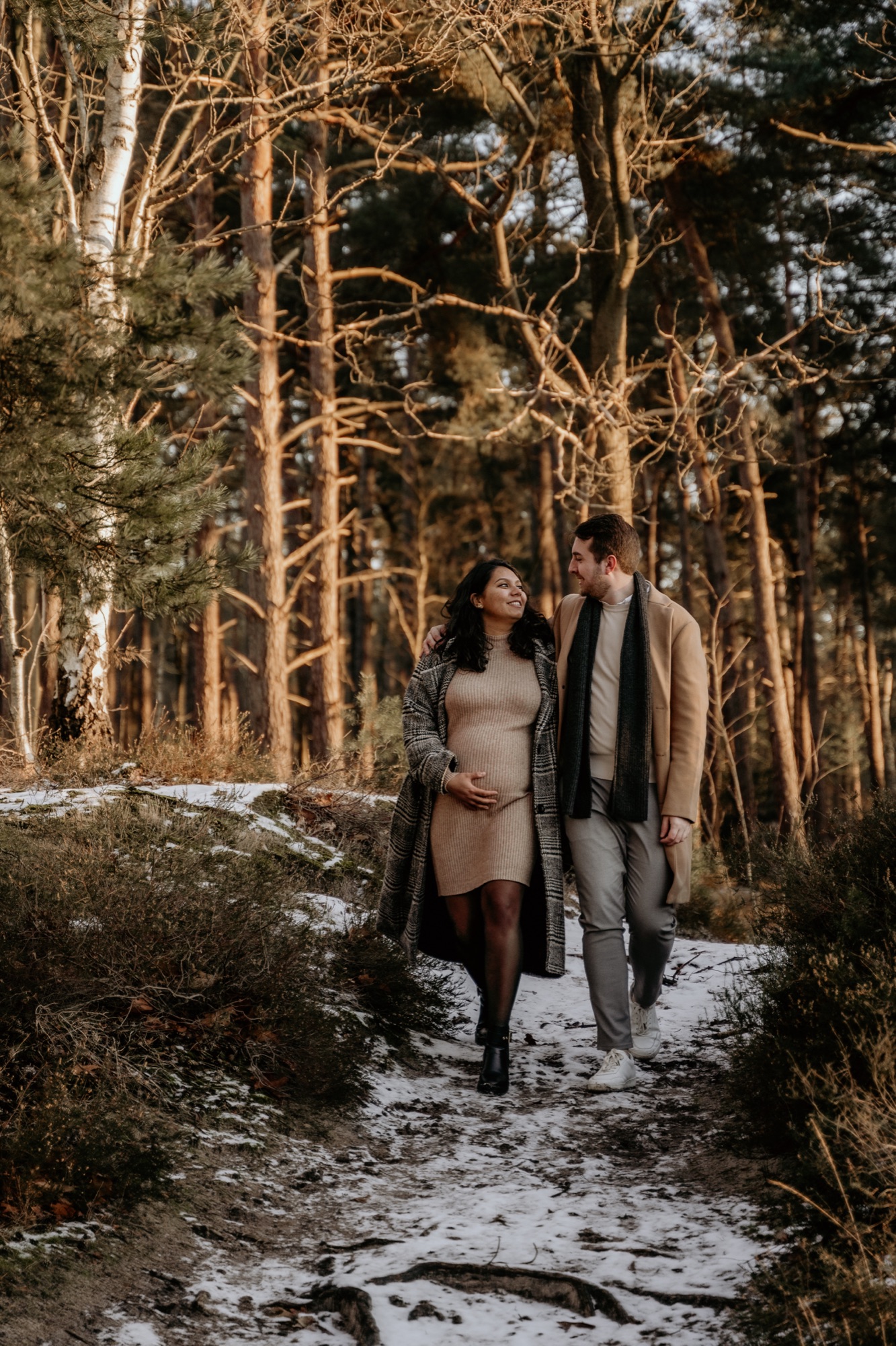 Koppel wandelt hand in hand over een besneeuwd bospad in warm avondlicht.