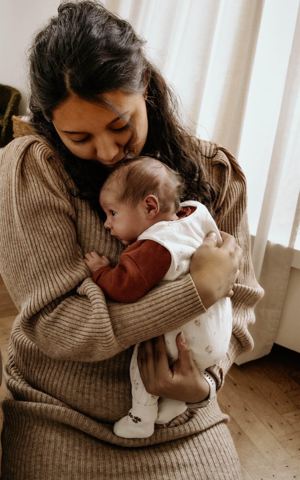 Moeder houdt haar pasgeboren baby teder vast bij een raam met zacht daglicht.